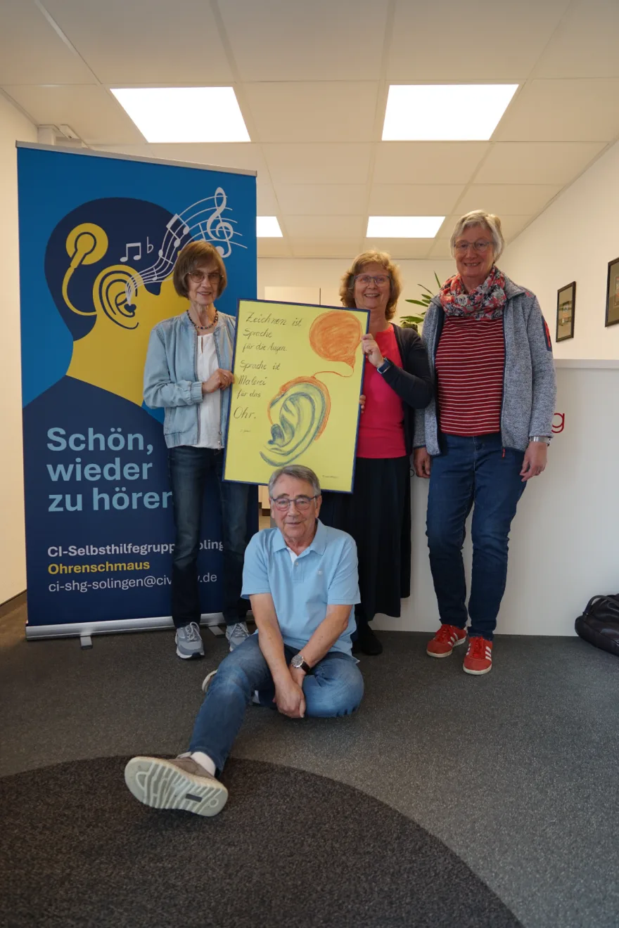 Vier Mitglieder der CI-Selbsthilfegruppe Ohrenschmaus stehen mit einem Plakat vor dem Rollup der Gruppe