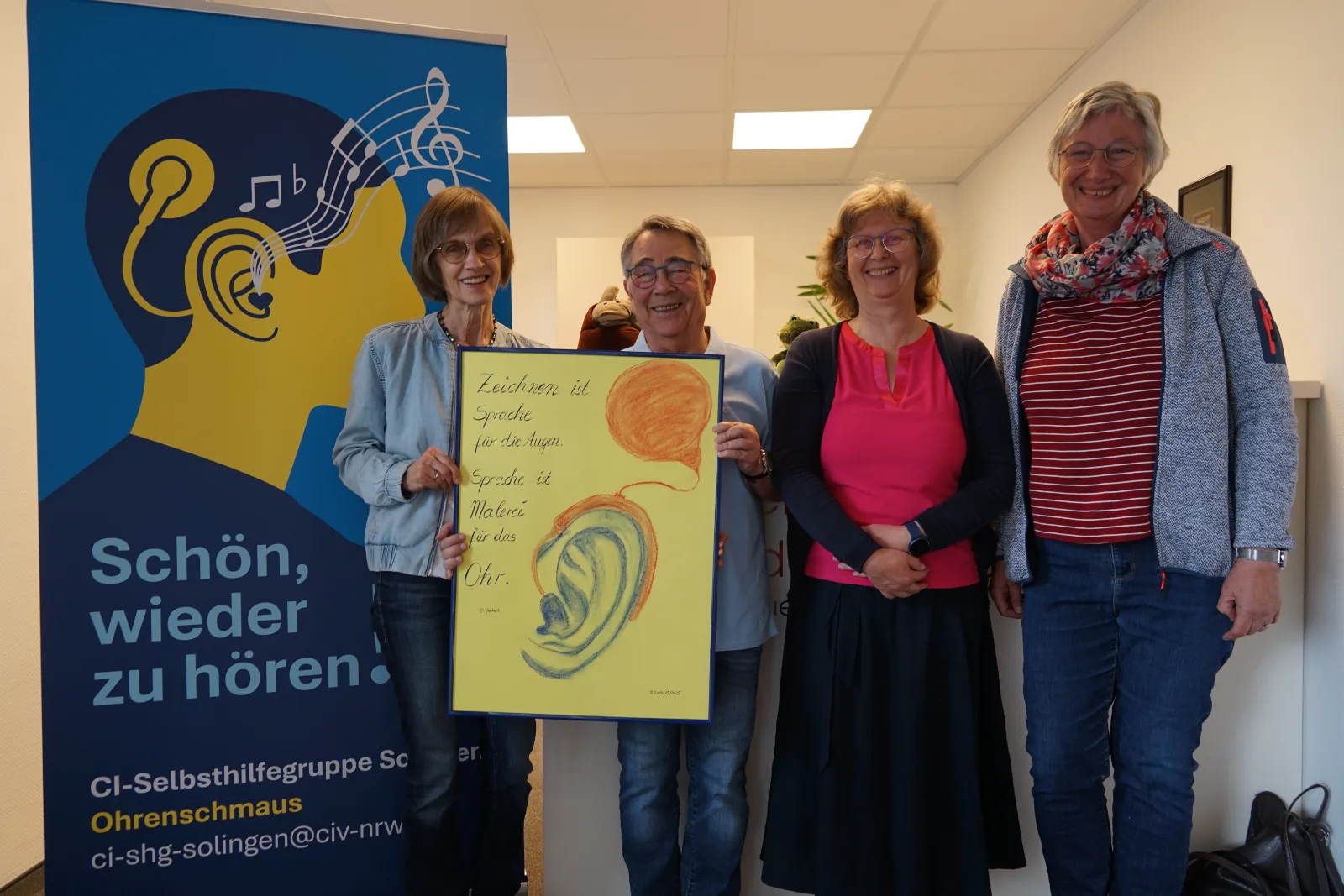Vier Mitglieder der Selbsthilfegruppe stehen nebeneinander und halten ein gemaltes Plakat mit einem Ohr